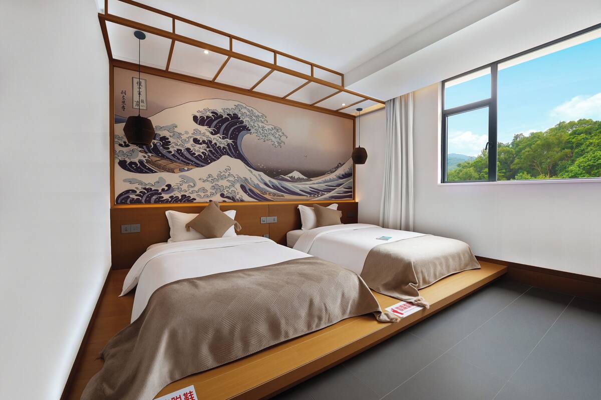A hotel room with two beds, a large wave painting on the wall, a window with a view of trees, and a wooden platform.