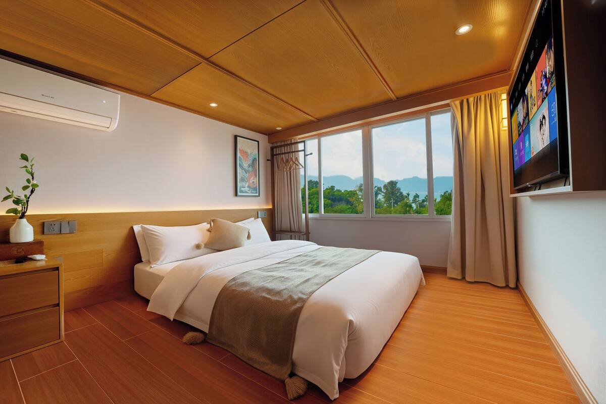 A hotel room with a large bed, wooden flooring, a mounted TV, and a window with a view of the outdoors.