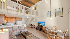 A spacious living area with a high ceiling, a balcony, a kitchen, and a dining area with wicker furniture.
