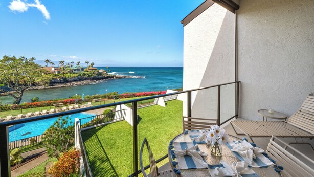 A balcony with a view of a pool and ocean, a table with a vase of flowers, and a lounge chair.