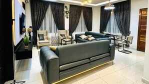 A modern living room with a black sofa, a dining area, and a TV mounted on the wall. - Mankatiwa Court Apartmentnt (Weija)