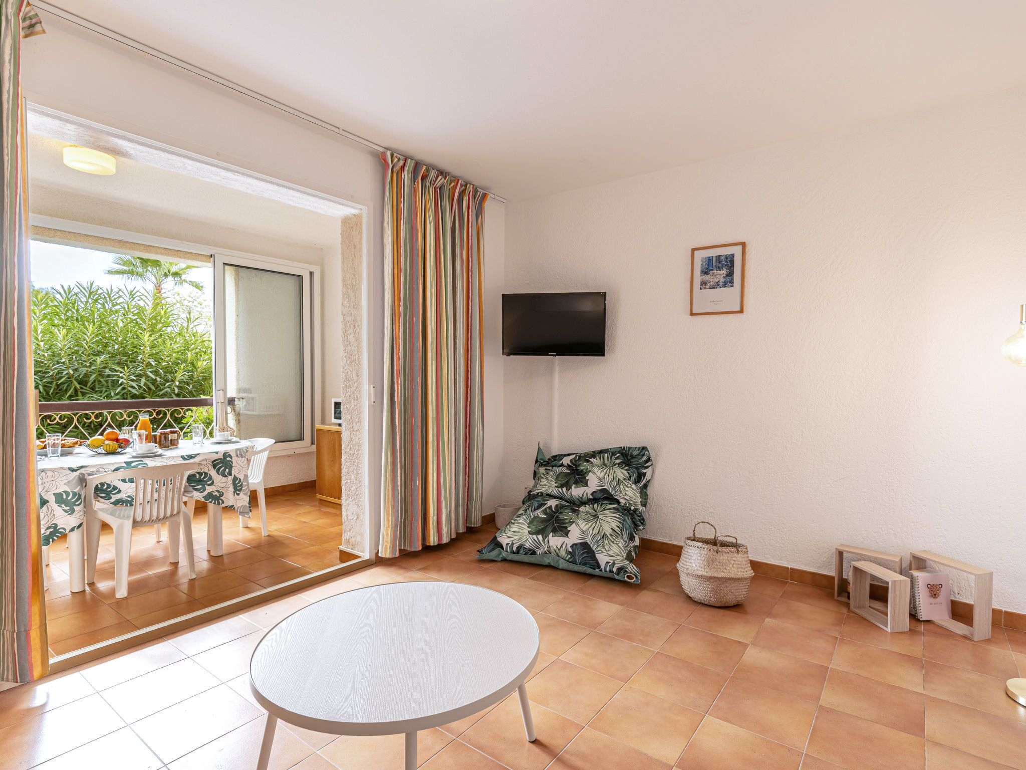 Una habitació amb balcó, taula de dinar amb cadires, televisió penjada a la paret i una taula rodona petita al centre.