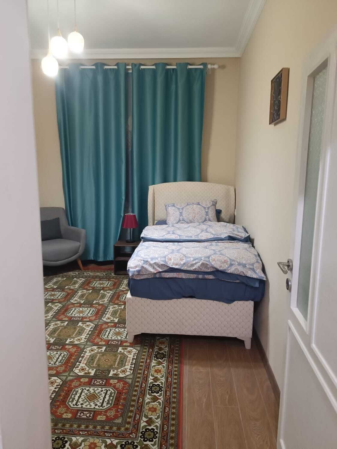 Une petite chambre à coucher avec un lit, une chaise, un tapis à motifs et des rideaux de couleur turquoise.
