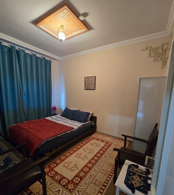 Ein Schlafzimmer mit einem Bett, einem Nachttisch, einem Sessel, einem Schreibtisch, einer Lampe, einer Deckenleuchte, einer Wanddekoration und einem Kleiderschrank.