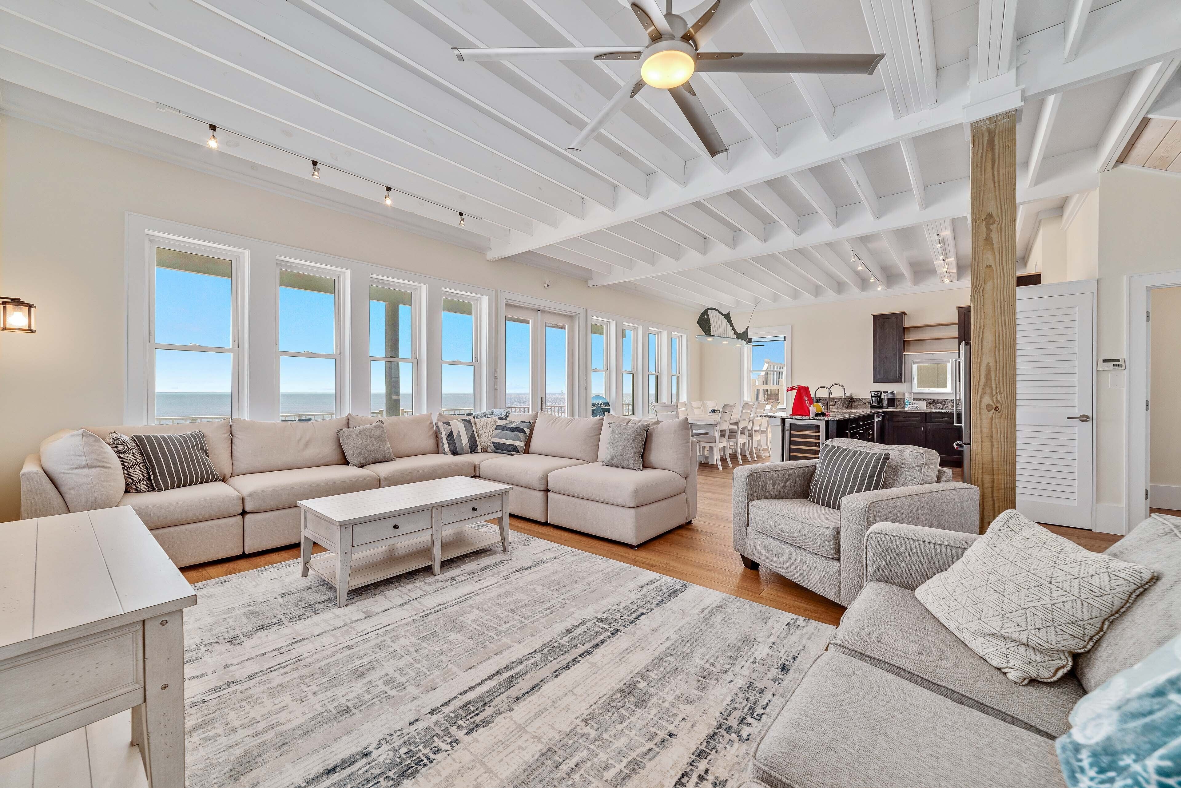 A spacious living room with a large sectional sofa, a coffee table, and a fireplace. There are multiple windows providing natural light and a view of the outdoors.