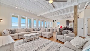 A spacious living room with a large sectional sofa, a coffee table, and a fireplace. There are multiple windows providing natural light and a view of the outdoors.