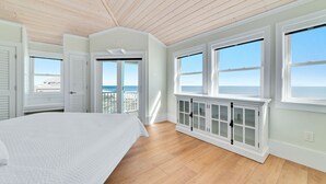 A bedroom with a large bed, wooden flooring, white walls, and a view of the ocean through large windows.