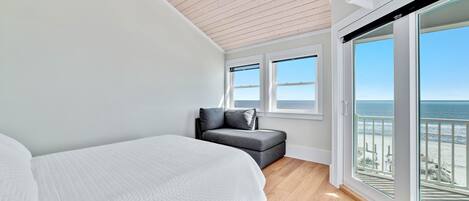 A bedroom with a bed, a gray sofa, large windows, and a view of the beach.