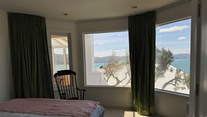A bedroom with a bed, a chair, a large window with a view of the sea and mountains, and green curtains. - Super large seaview house (Wellington)