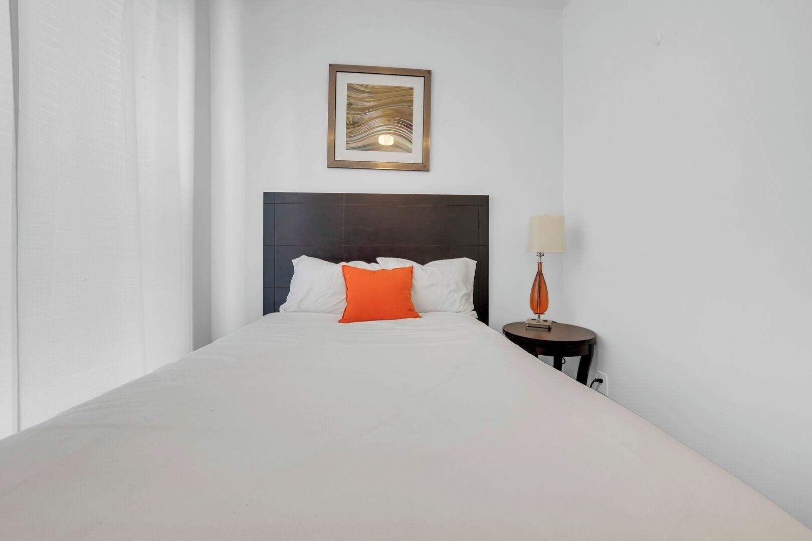 Une chambre d’hôtel avec un grand lit, une tête de lit sombre, du linge de lit blanc, un coussin décoratif orange, une œuvre d’art encadrée au mur et une petite table de chevet avec une lampe.