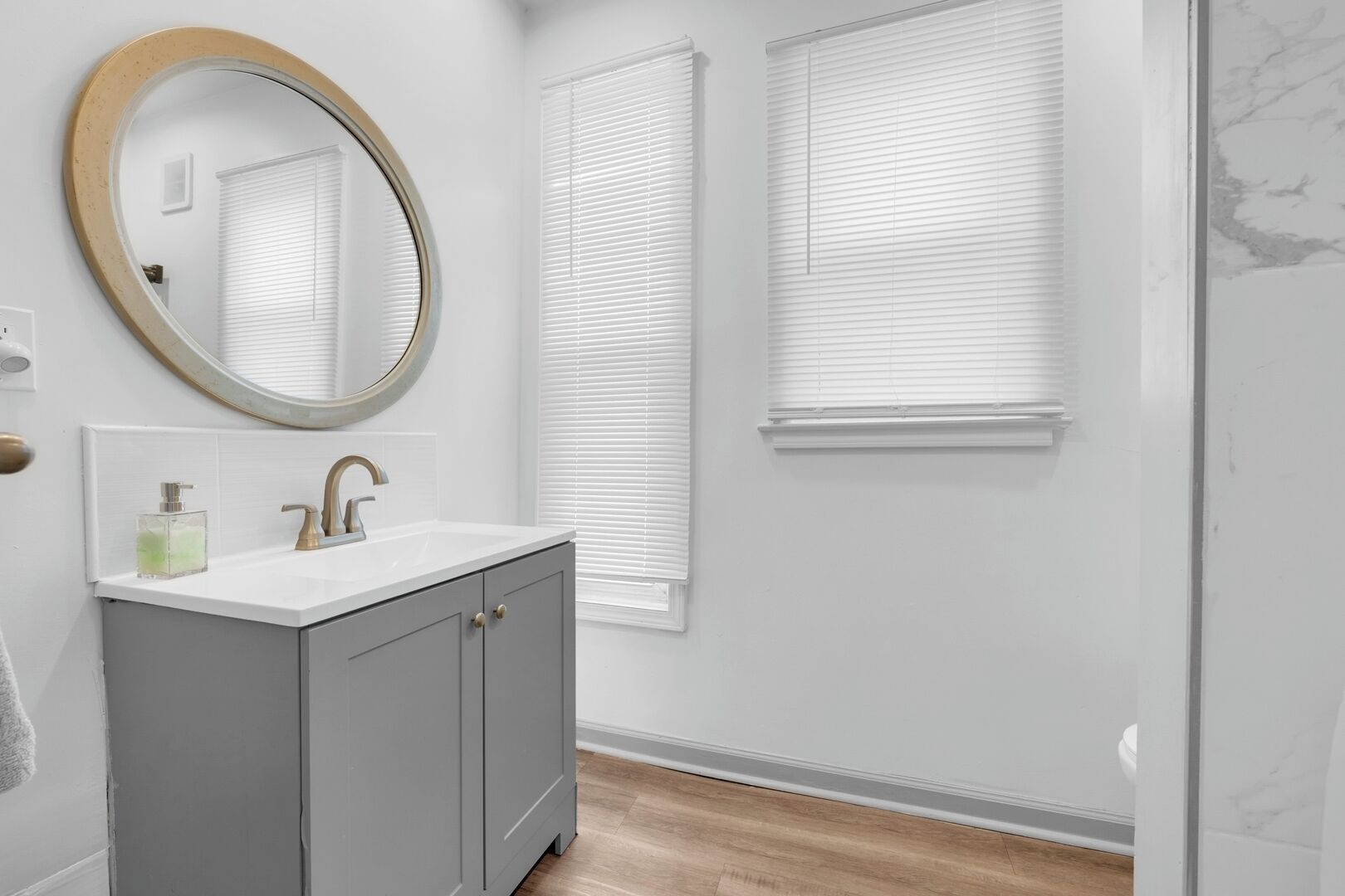 Een badkamer met een witte wastafel, grijze kastjes, een ronde spiegel en witte jaloezieën.