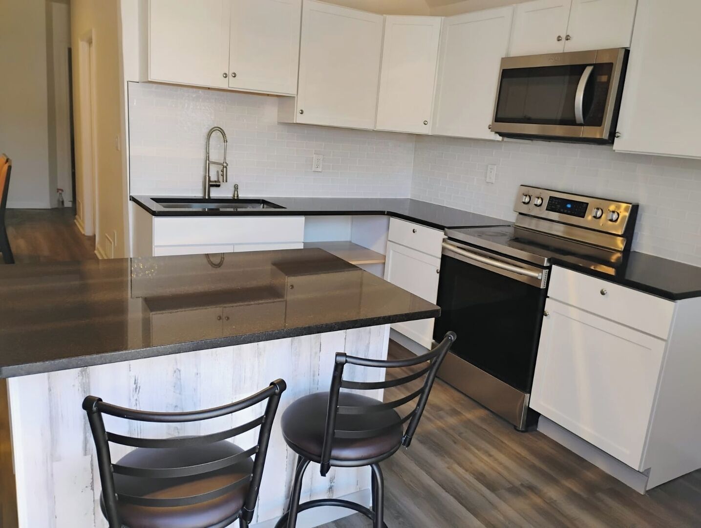 A modern kitchen with white cabinets, a stainless steel microwave, an oven, and a granite countertop. There are two bar stools with metal frames.