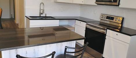 A modern kitchen with white cabinets, a stainless steel microwave, an oven, and a granite countertop. There are two bar stools with metal frames.