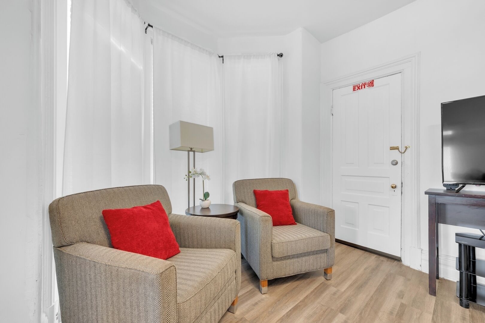 A room with two armchairs, a small table with a lamp, a television on a stand, and a white door with a red sign.