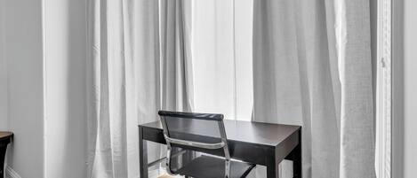 Un espacio de oficina moderno con un escritorio negro y una silla de oficina, paredes blancas y cortinas translúcidas.