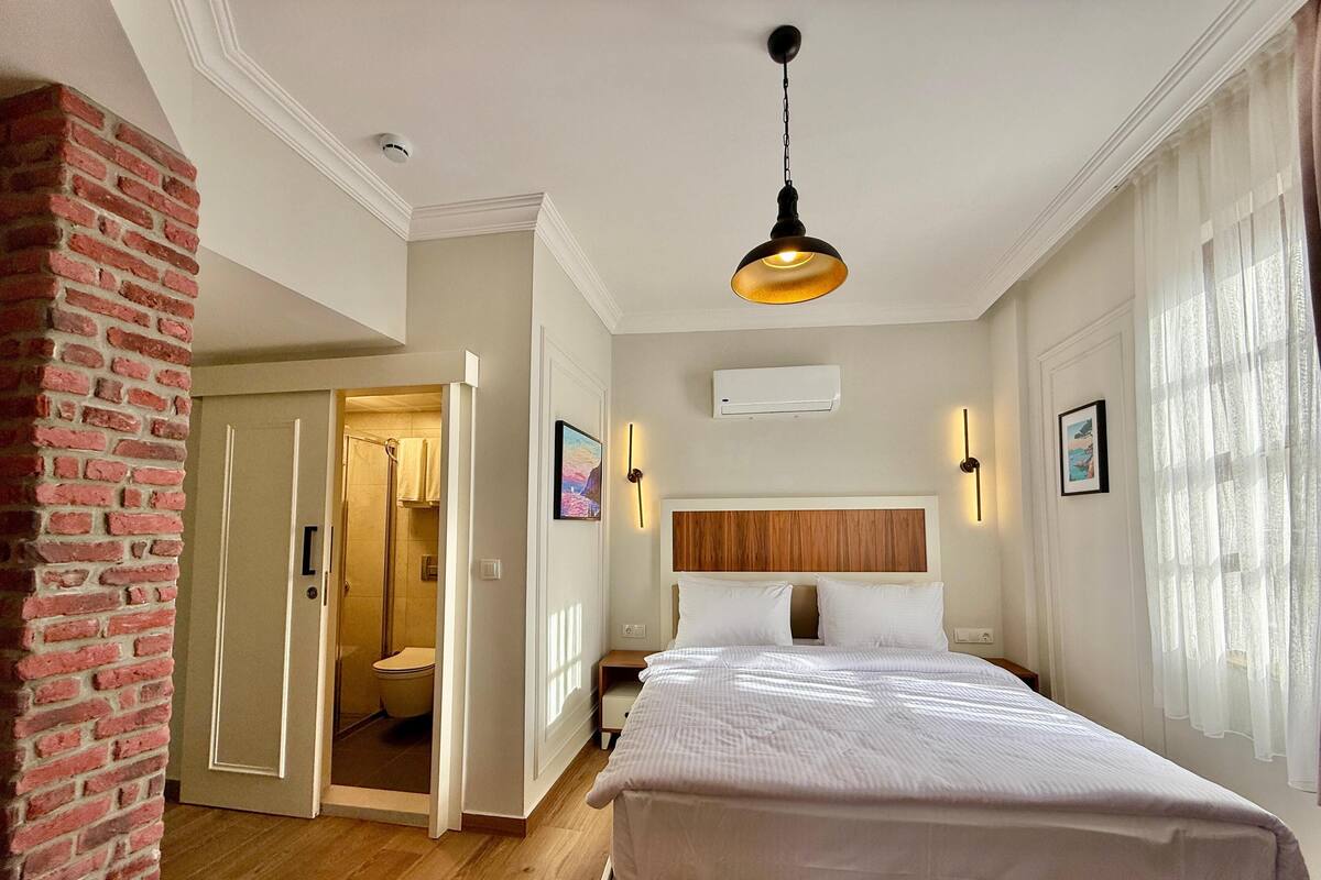 A hotel room with a wooden headboard, a large bed, a hanging light fixture, a bathroom with a glass door, and a brick wall.
