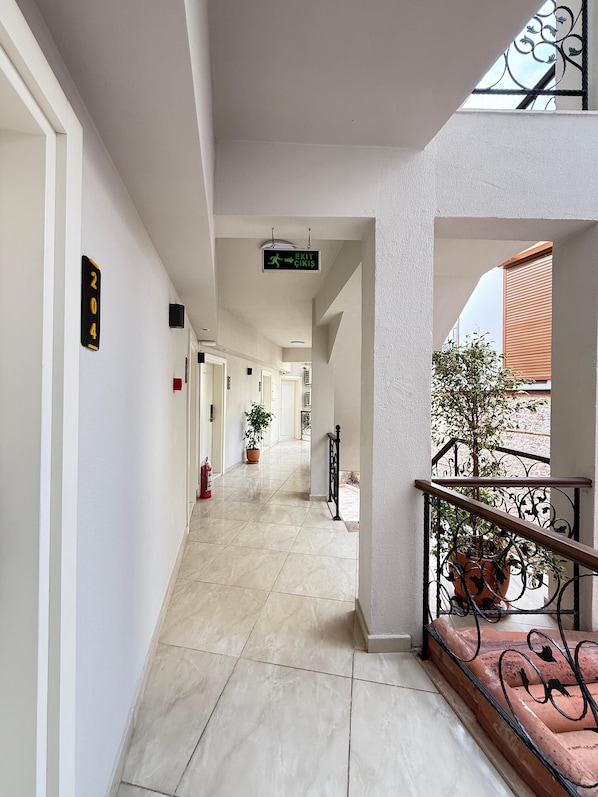 A corridor with a tiled floor, a fire extinguisher, a potted plant, and a balcony with a wrought iron railing.