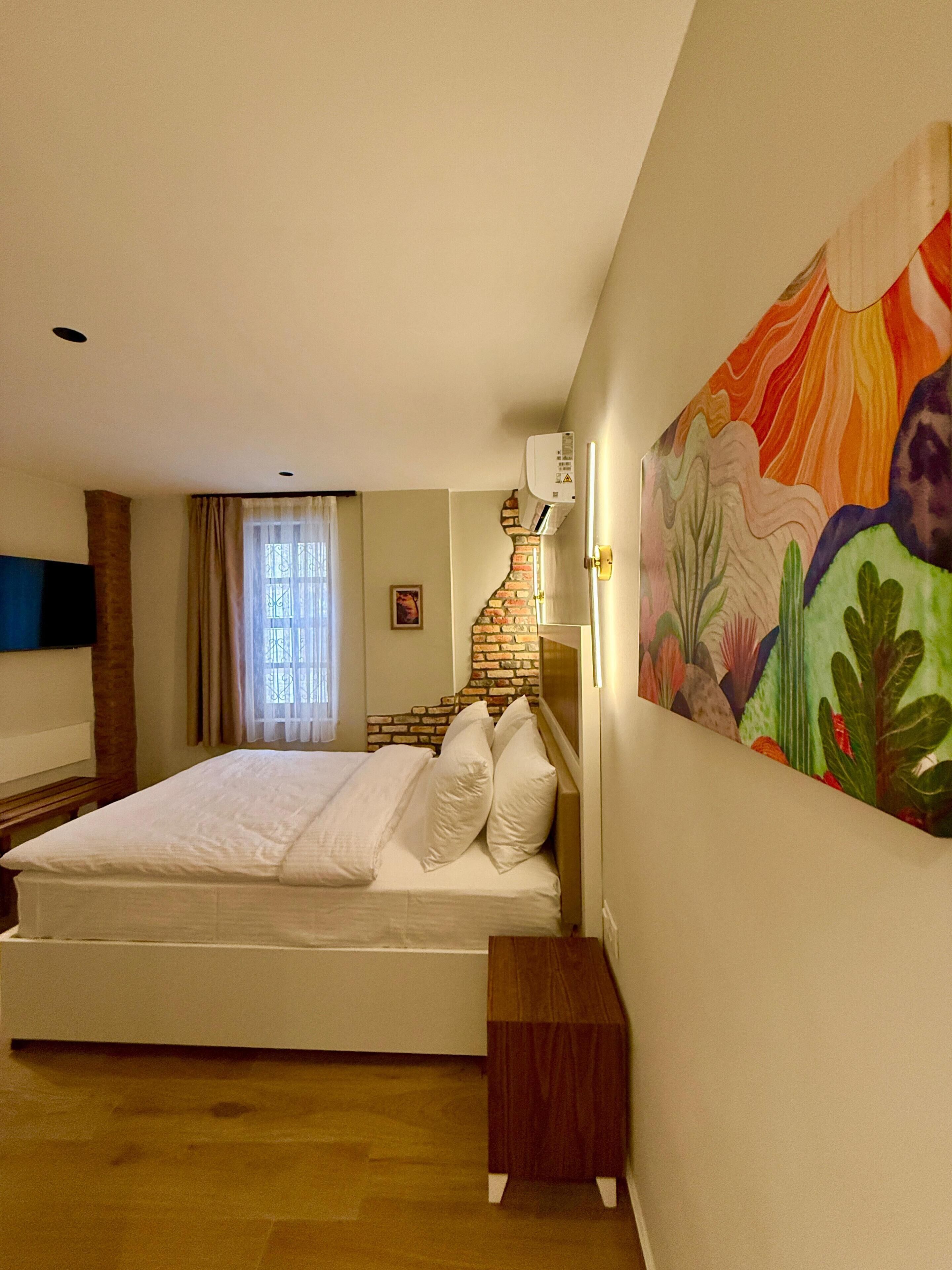 A bedroom with a bed, a wooden bench, a wall-mounted air conditioner, and a colorful painting.