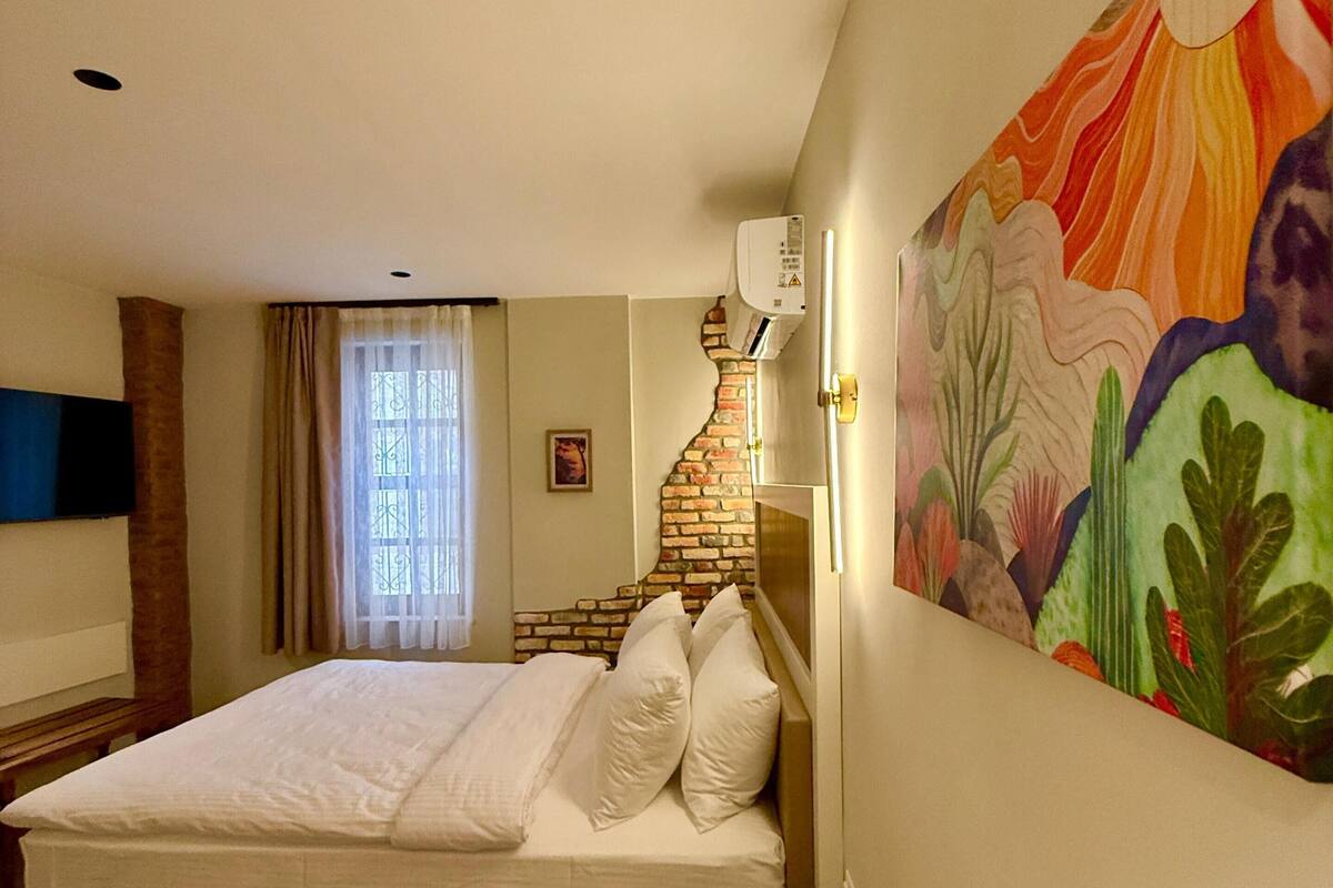 A bedroom with a bed, a wooden bench, a wall-mounted air conditioner, and a colorful painting.