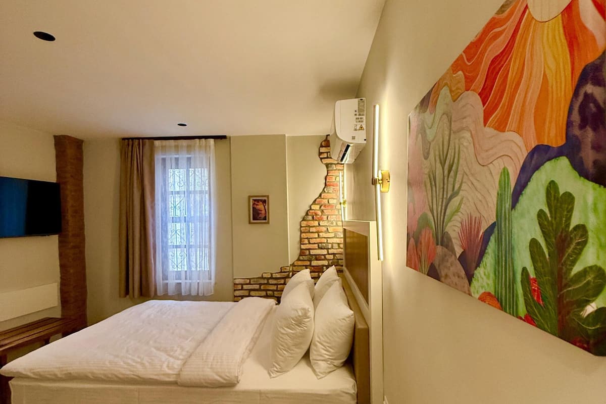 A bedroom with a bed, a wooden bench, a wall-mounted air conditioner, and a colorful painting.