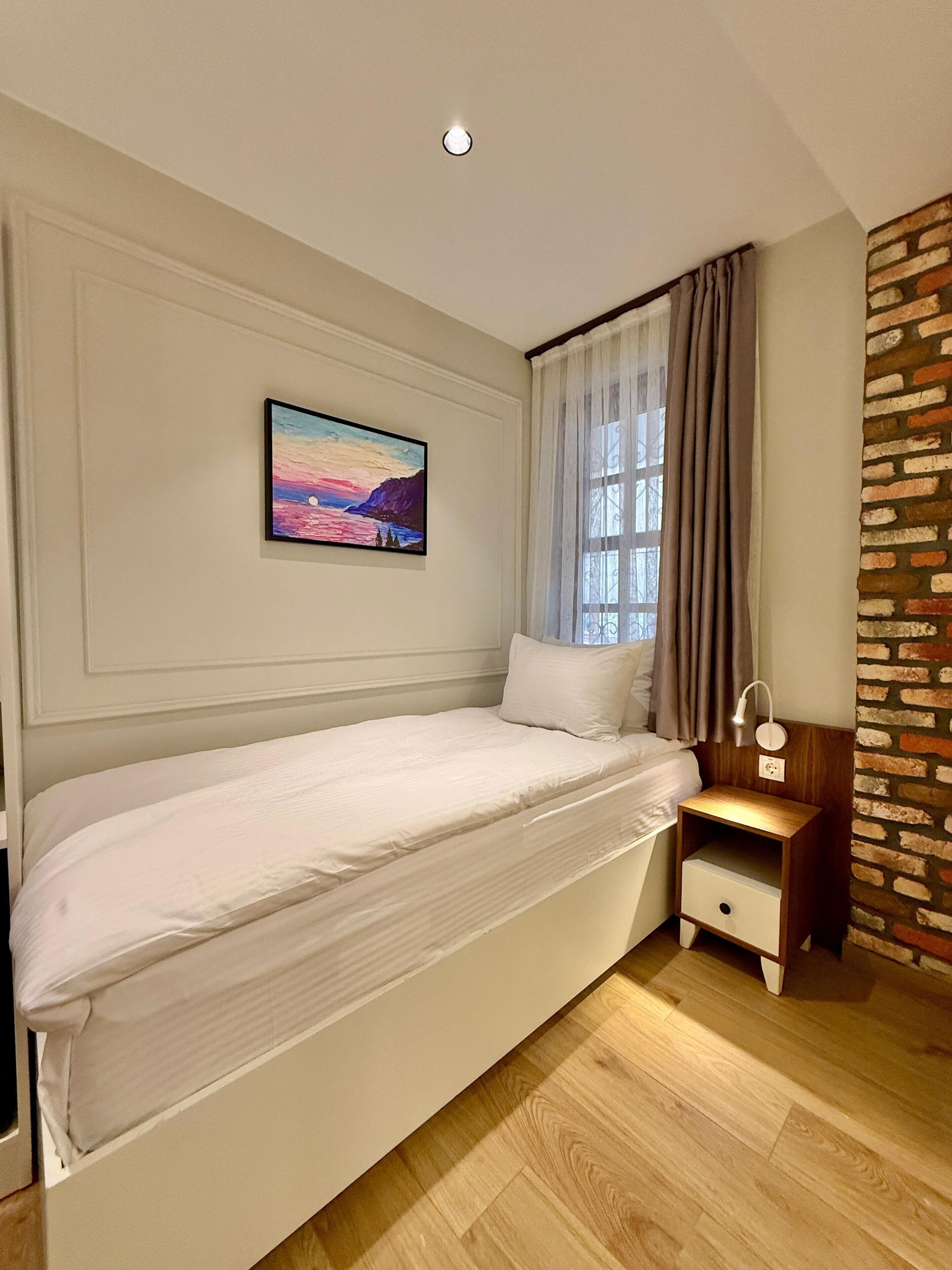 A bedroom with a bed, a nightstand, a lamp, a painting, and a window with curtains.