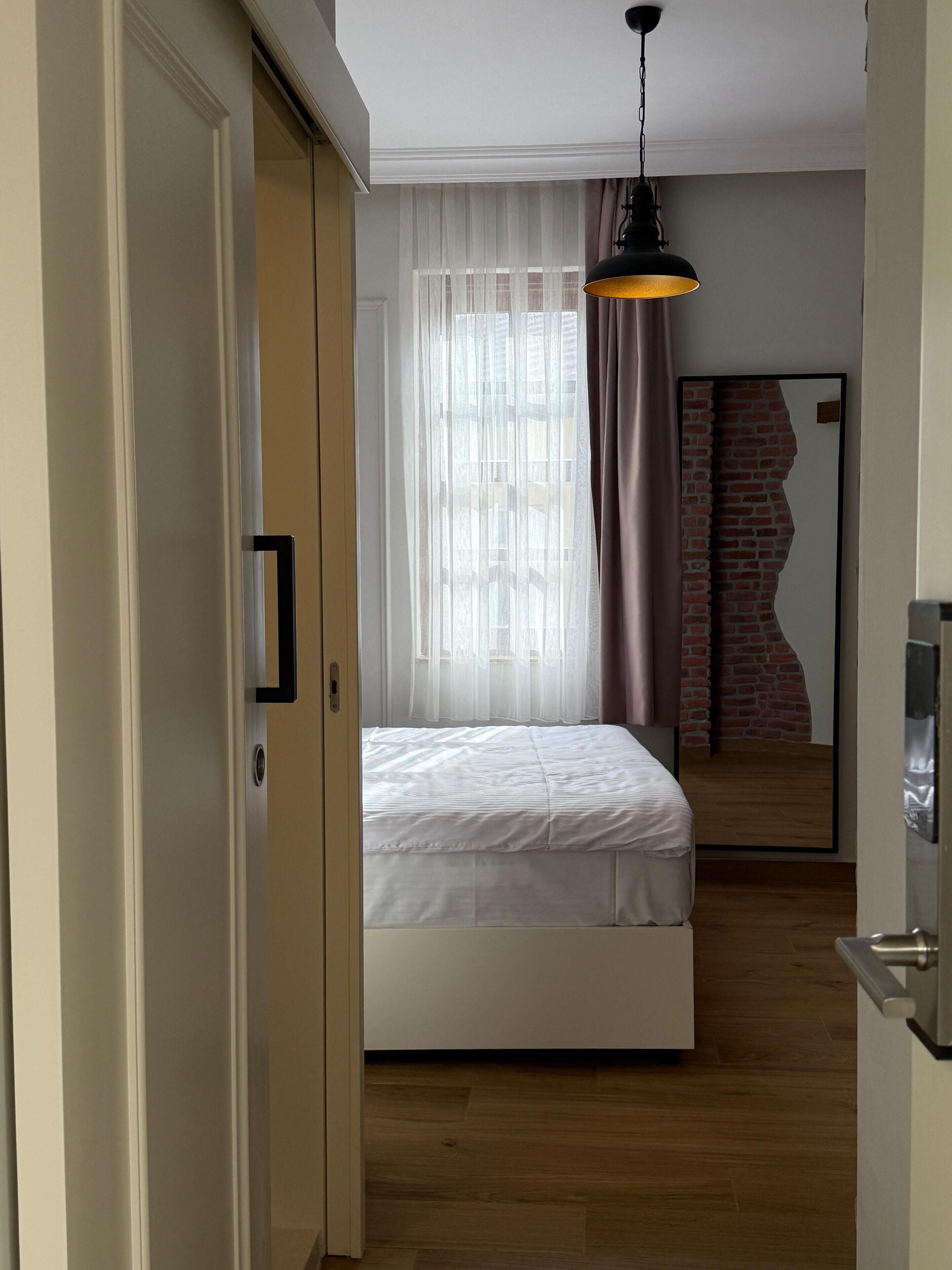 A bedroom with a bed, a wooden floor, a window with sheer curtains, a hanging light fixture, and a mirror reflecting a brick wall.