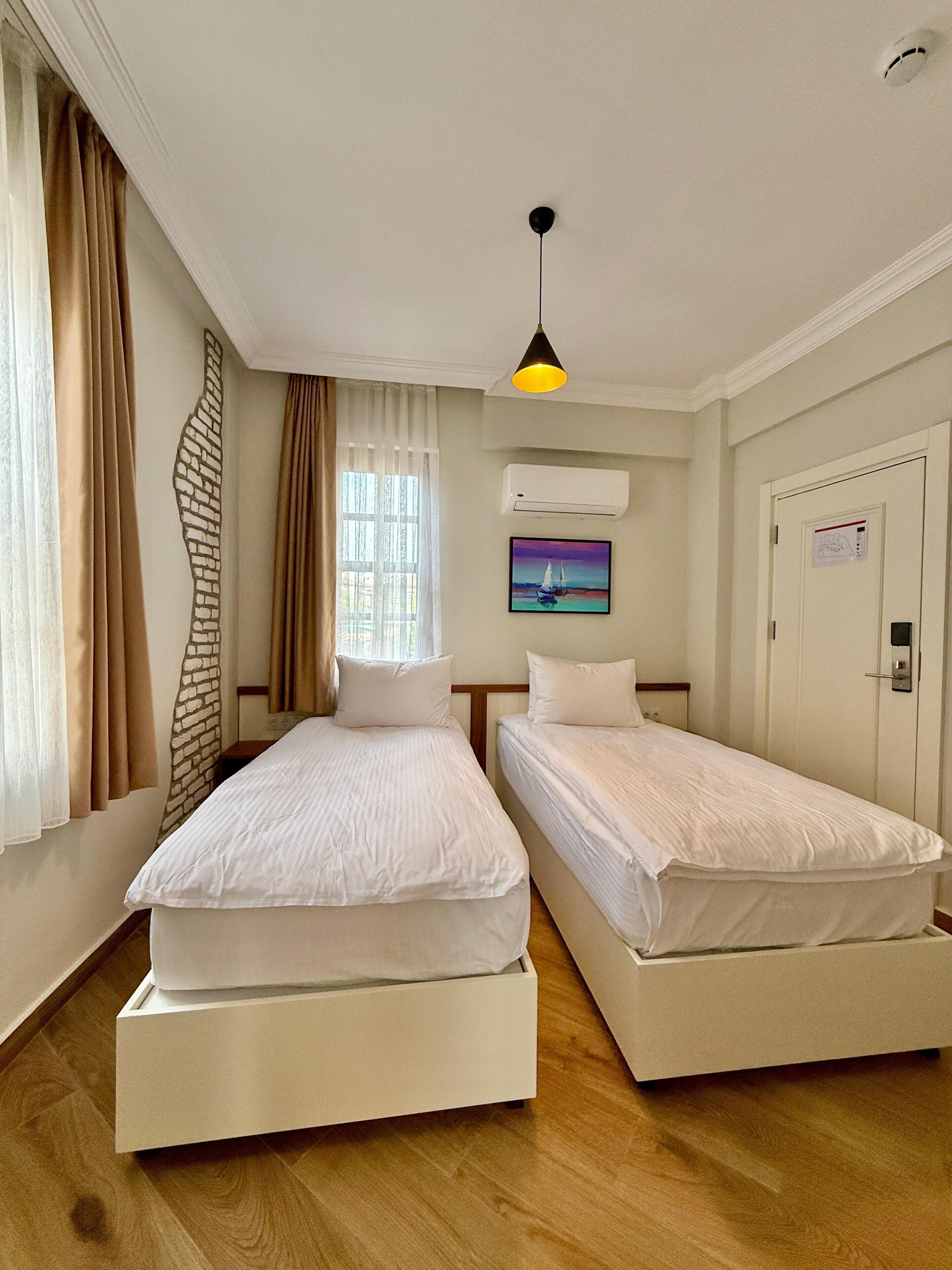 Ein Hotelzimmer mit zwei Einzelbetten, einem Fenster mit Vorhängen, einer Klimaanlage, einem Gemälde und einer Tür mit Schloss.