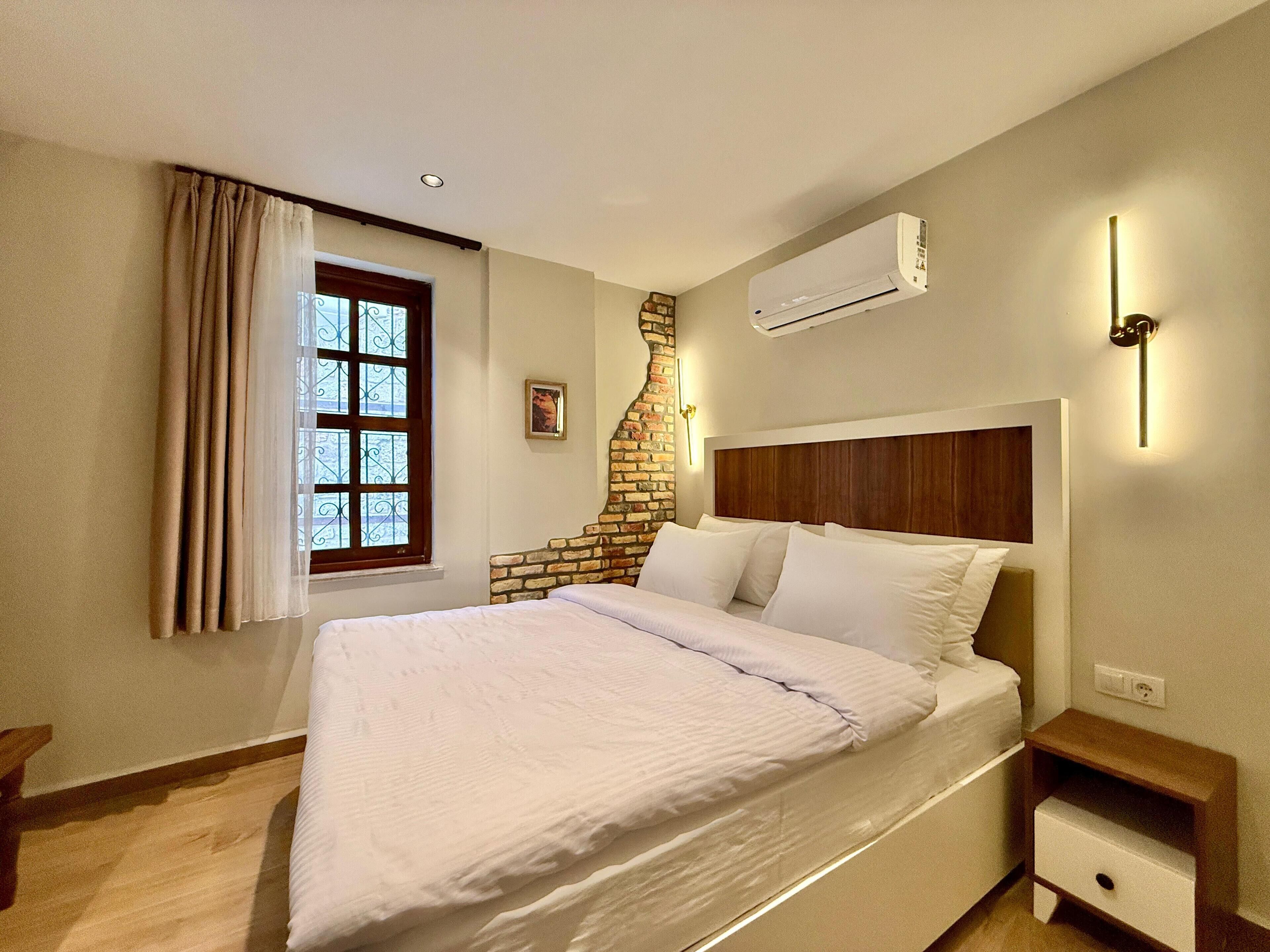 Quarto com uma cama grande, parede de destaque em tijolo, janela com cortinas, ar condicionado e mesa de cabeceira com gaveta.
