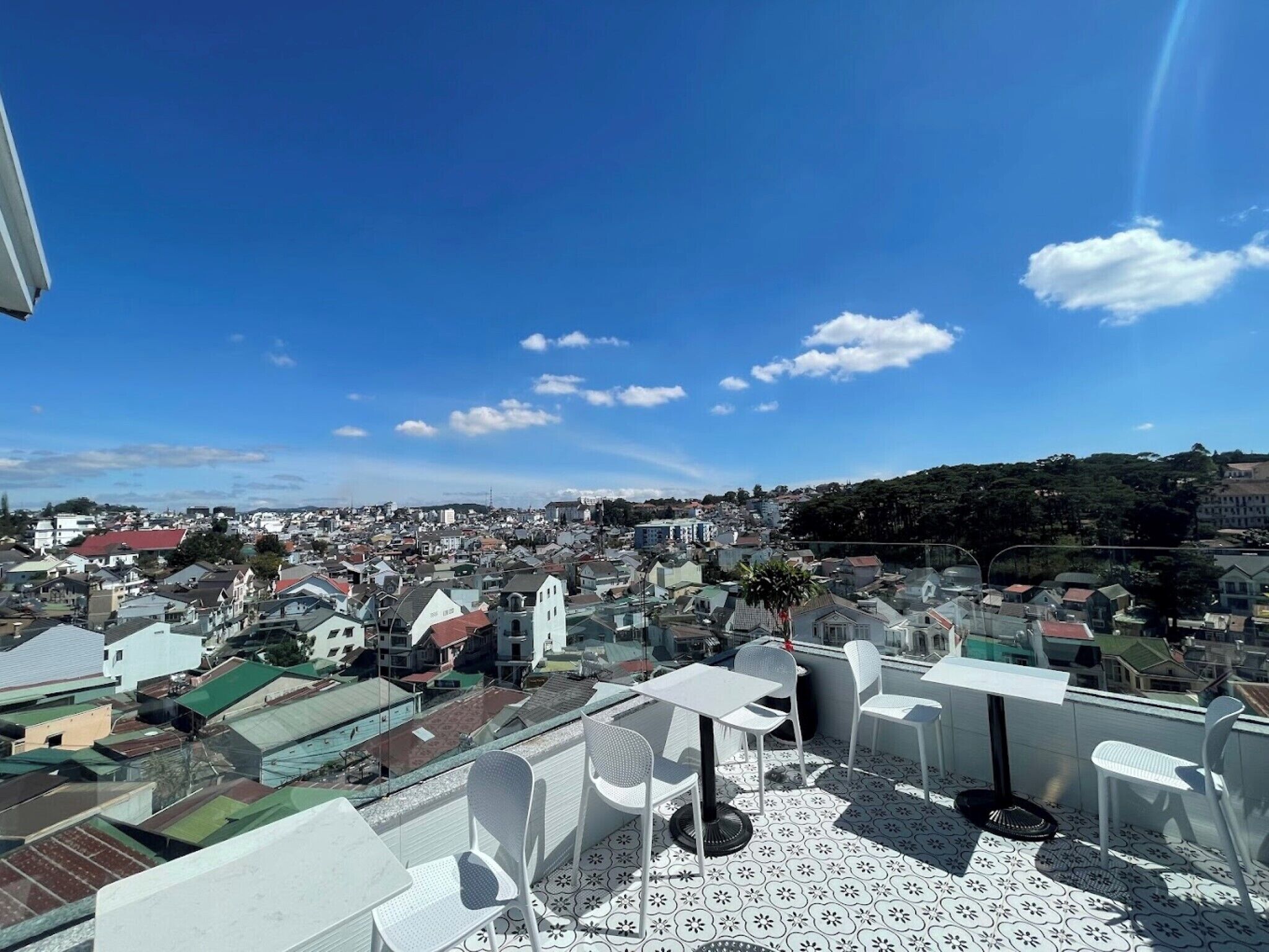 A rooftop terrace with white chairs and tables, offering a view of a densely populated town and hills in the distance.