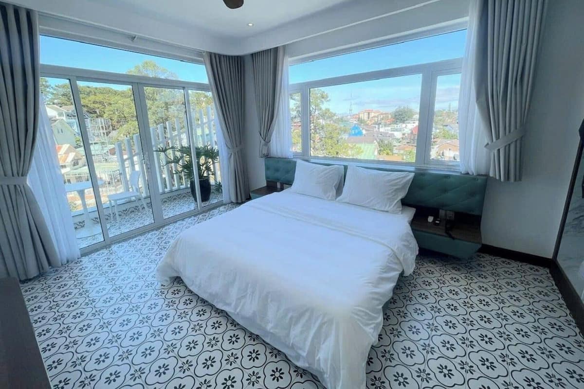 A bedroom with a large bed, a green upholstered bench, and a view of a residential area through large windows.