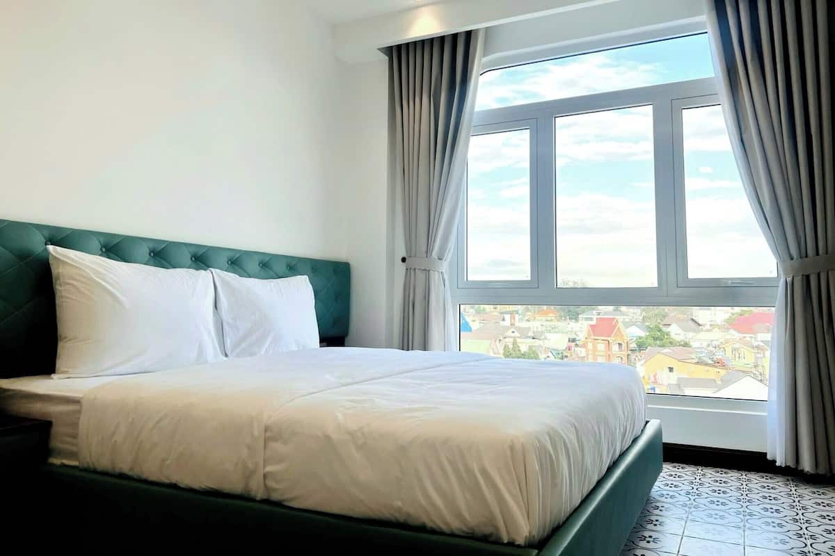 A bedroom with a bed, a view of a town through a large window, and a green headboard.