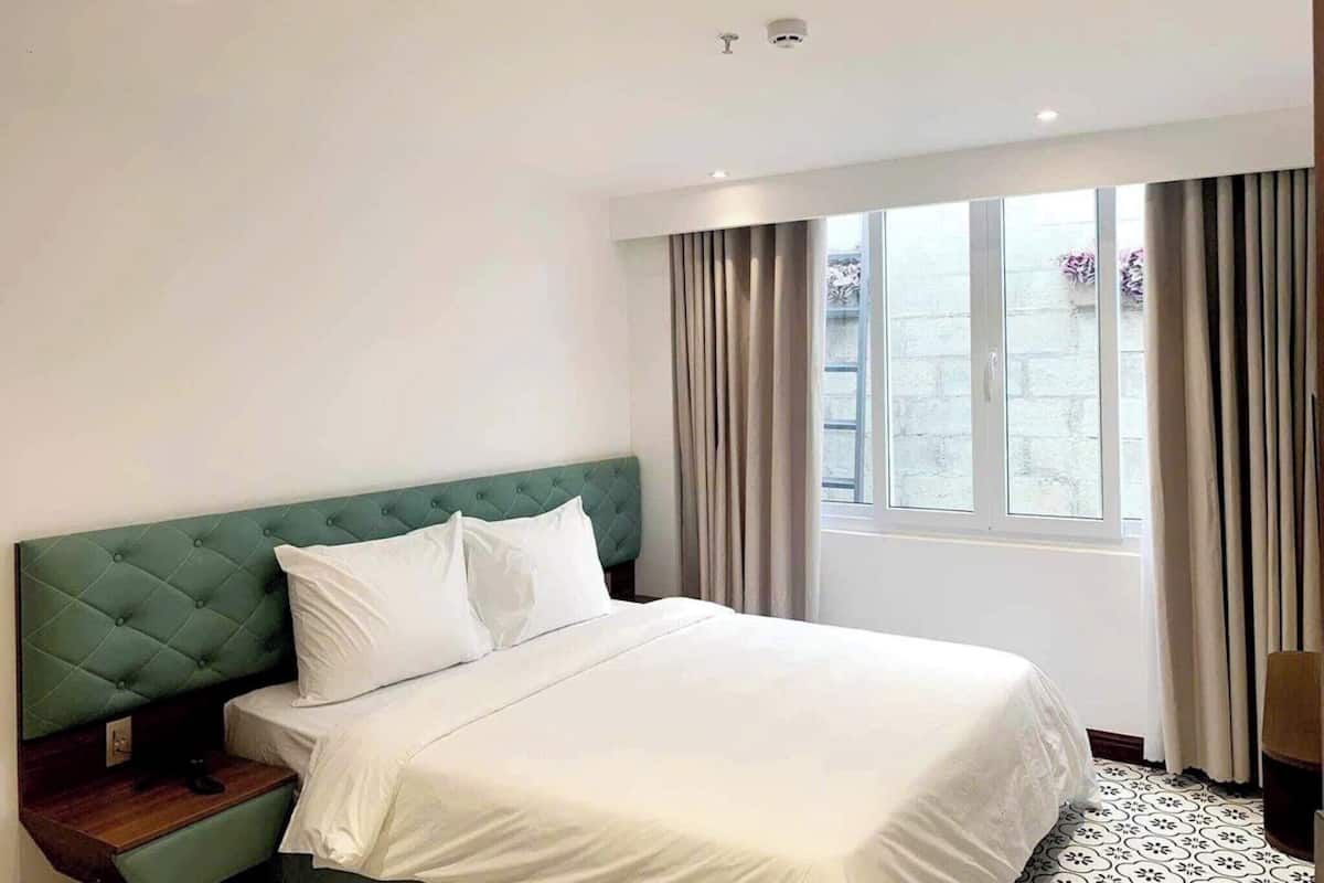 A hotel room with a green tufted headboard, white bedding, a wooden nightstand, and a large window with curtains.