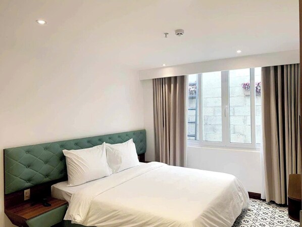 A hotel room with a green tufted headboard, white bedding, a wooden nightstand, and a large window with curtains. - Le's Dalat Sky Hotel  (Cam Ly - Da Lat)