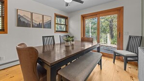 Ein Esszimmer mit Holztisch, Stühlen, einer Bank und einem Deckenventilator. Es gibt Fenster mit Blick auf Bäume und eine Tür, die nach draußen führt.
