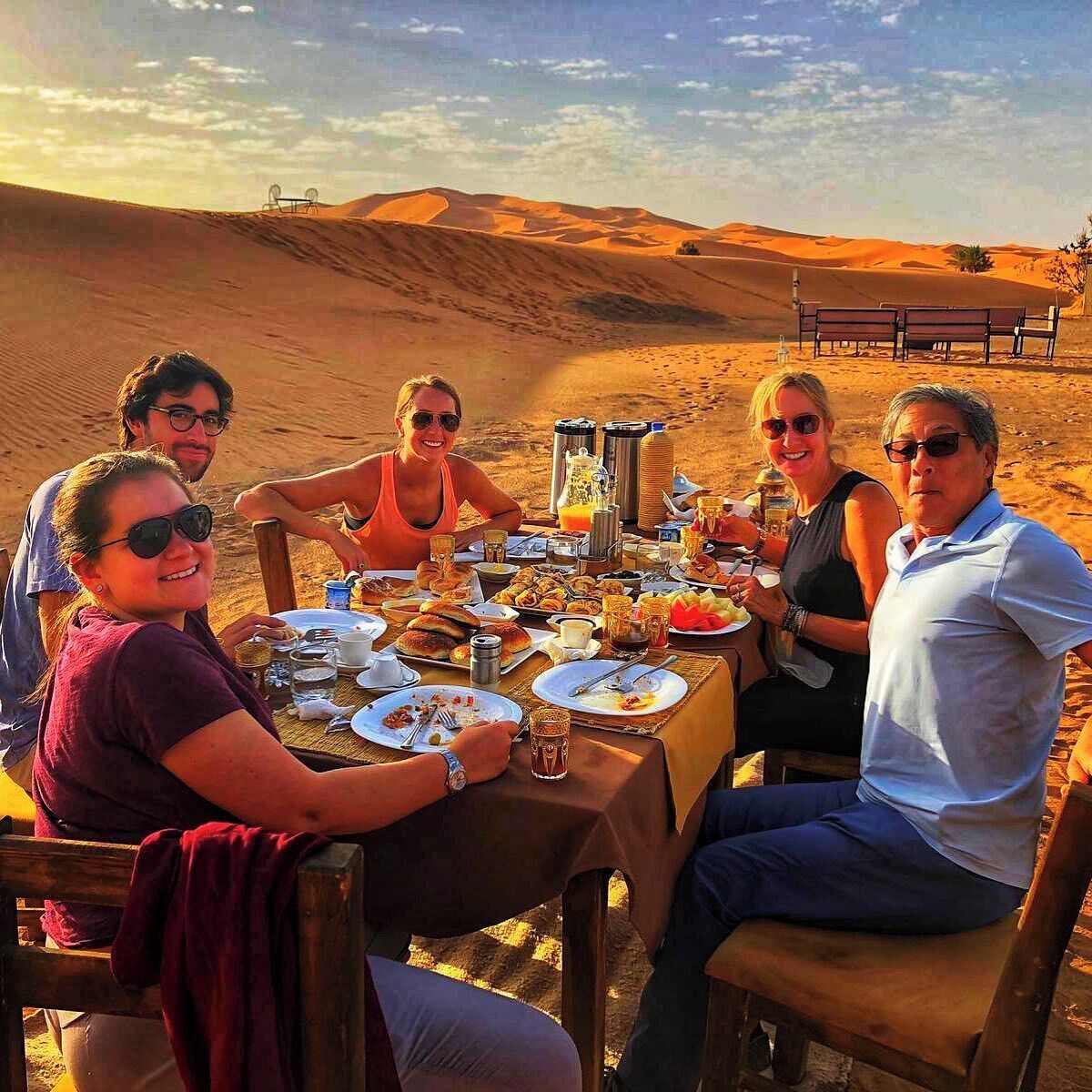 A group of people dining at a table set up in a desert with a view of sand dunes in the background.