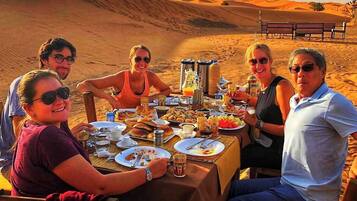 A group of people dining at a table set up in a desert with a view of sand dunes in the background.