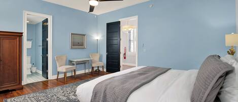 A bedroom with a bed, a ceiling fan, a chair, a small table, and a door leading to a bathroom.