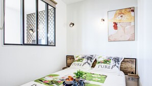 Een slaapkamer met een bed, een raam met uitzicht op een gebouw, een wandlamp en een lijst met een afbeelding.