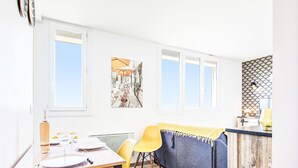Een lichte eetruimte met een houten tafel, gele stoelen en een grijze bank. Er zijn twee ramen met witte kaders, een aan de muur hangend schilderij en een behang met patroon achter de bank.
