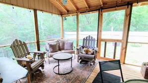 A covered porch with wooden furniture, including two Adirondack chairs and a sofa, a small round table, and a view of a wooded area through large windows. - Charming NEW 2 BR Cottage, WiFi & Pet Friendly in Convenient Steele (Steele)