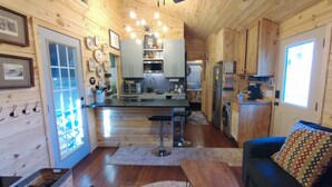 A cozy cabin interior with a wooden interior, a kitchen area with a central island, and a living room with a sofa and a patterned cushion. - Charming NEW 2 BR Cottage, WiFi & Pet Friendly in Convenient Steele (Steele)