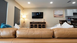 A living room with a brown leather sofa, a TV mounted on the wall, a lamp, and a kitchen area with cabinets and a countertop. - Stunning Pet Friendly House with King Bed and Texas Size Backyard in Weslaco (Weslaco)