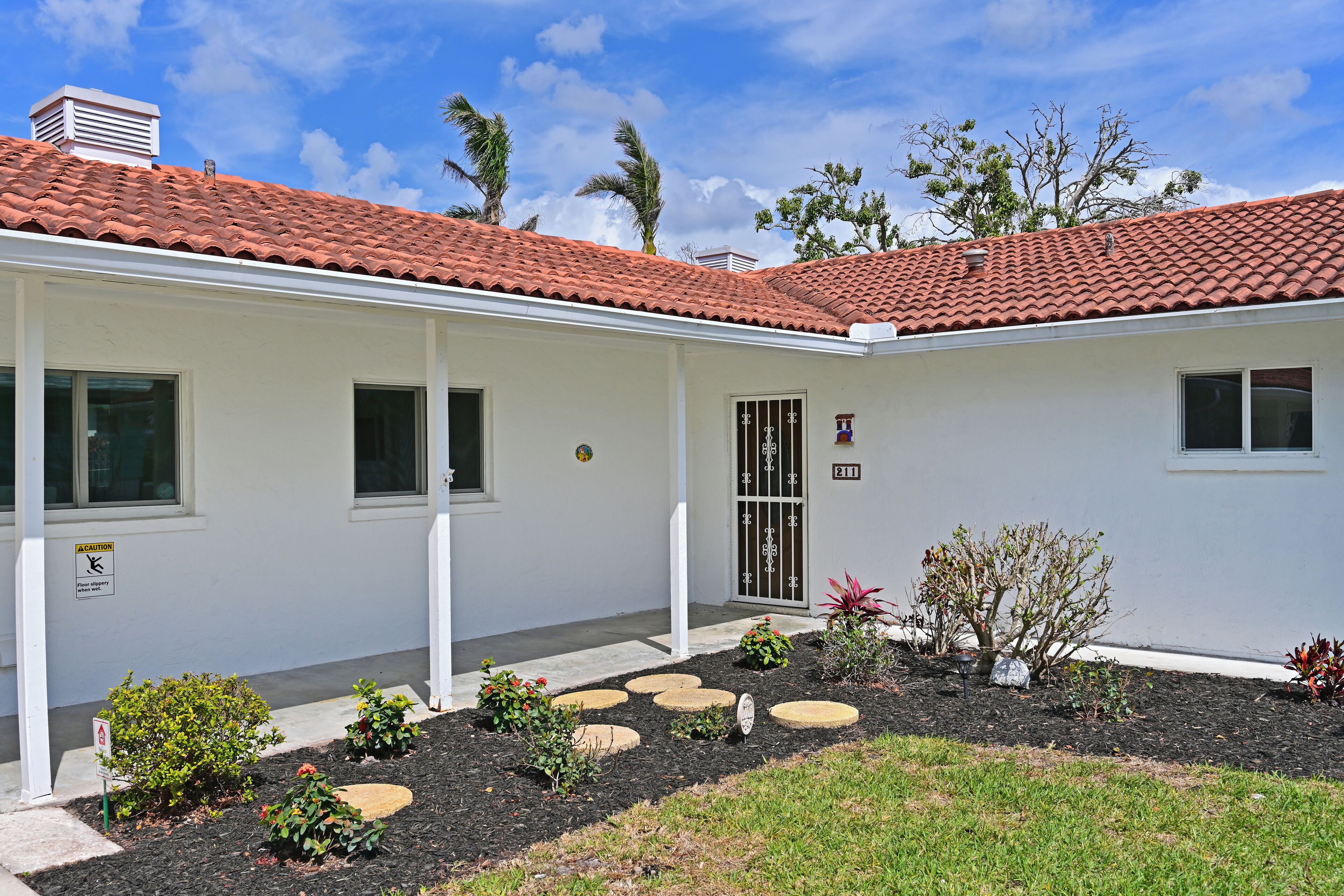 A white building with a red-tiled roof, a covered porch, and a small garden with stepping stones and plants.