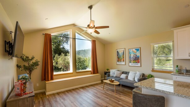 A spacious living area with a ceiling fan, large windows, a sofa, and a kitchen counter. - New 1 Bed Near Yosemite, Fire Pit, Washer, Dryer (Mariposa)
