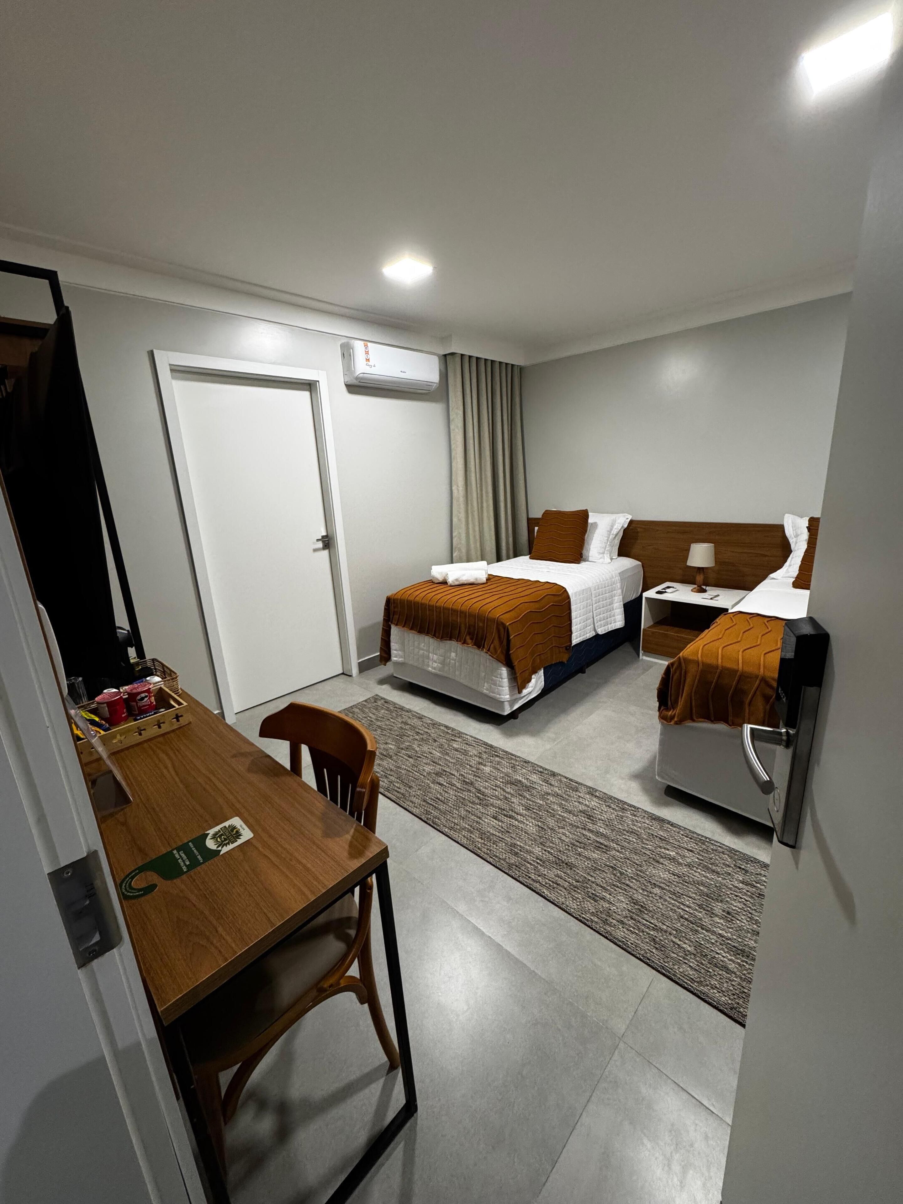 A hotel room with two beds, a desk, and a chair. There is a door, an air conditioning unit, and a small table with a basket of items.
