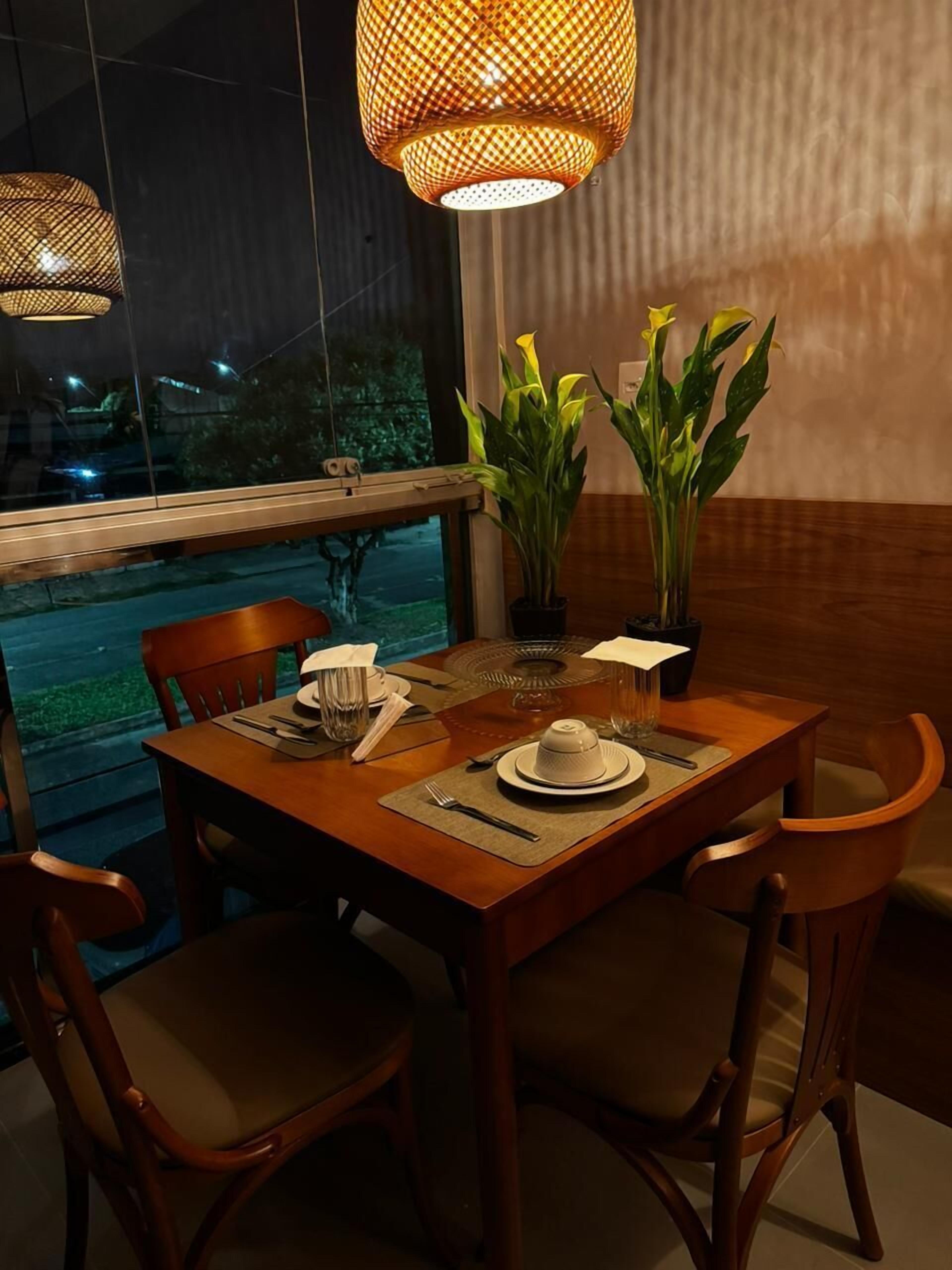 Una mesa de comedor dispuesta con platos, cubiertos y una tetera, rodeada de sillas, plantas en macetas y vistas al exterior.