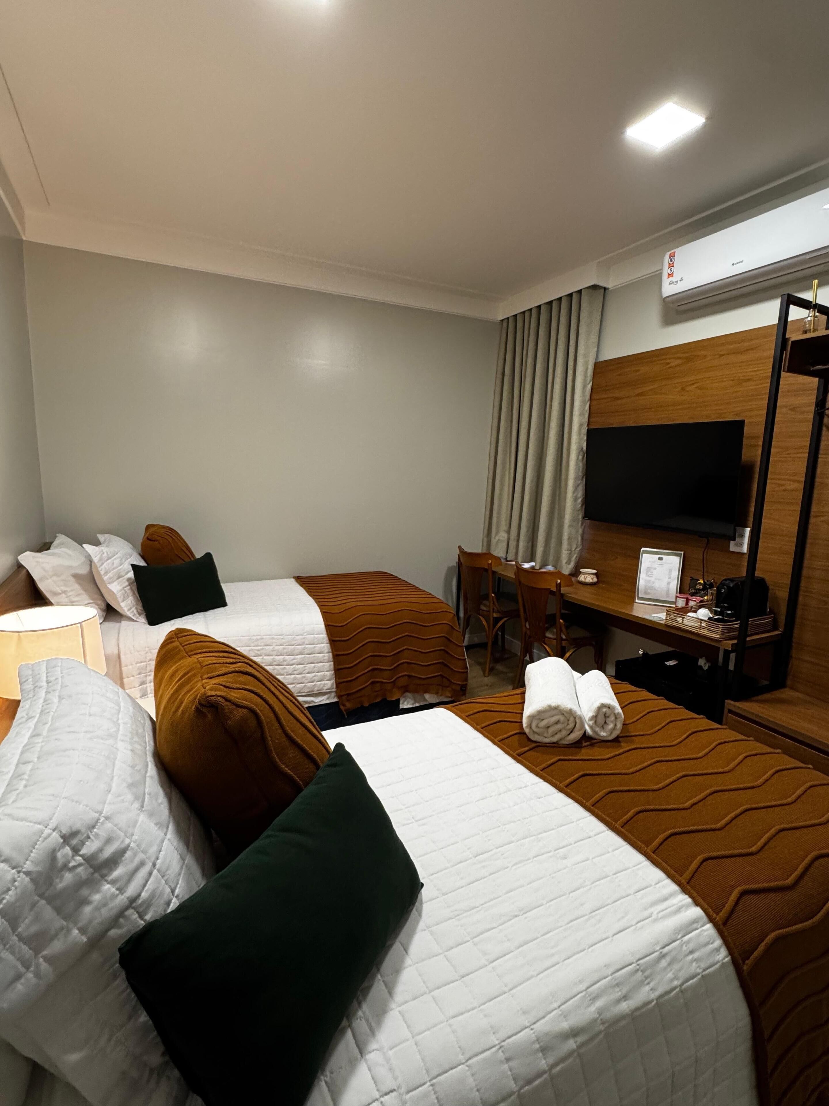A hotel room with two beds, a desk, a chair, a television, and an air conditioning unit.