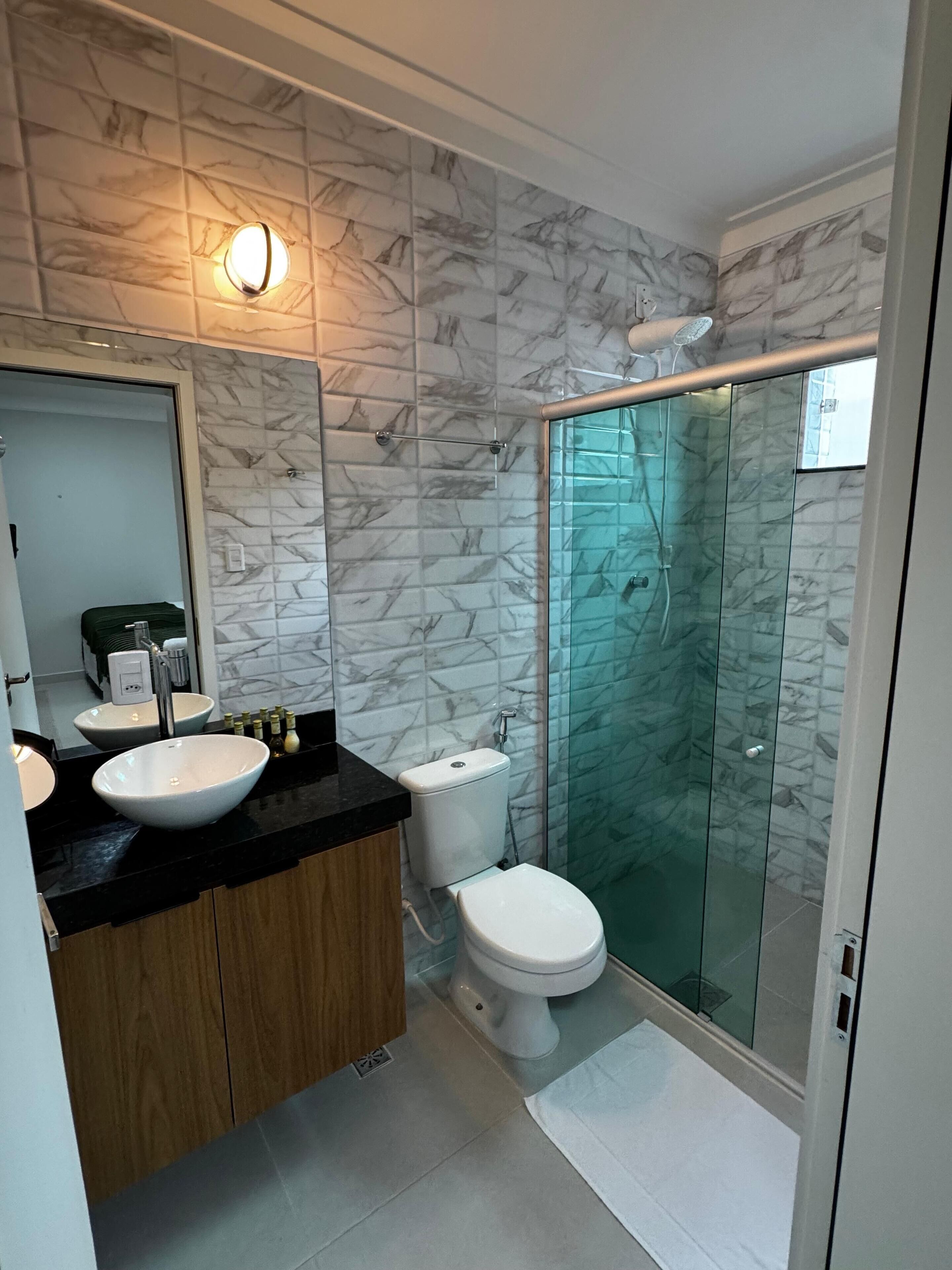 A modern bathroom with marble-patterned walls, a black countertop, wooden cabinets, a white sink, a toilet, and a glass-enclosed shower.