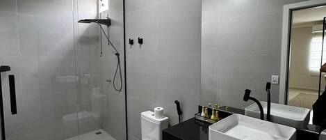 Un baño moderno con ducha envuelta en vidrio, inodoro y dos lavamanos sobre encimera negra.