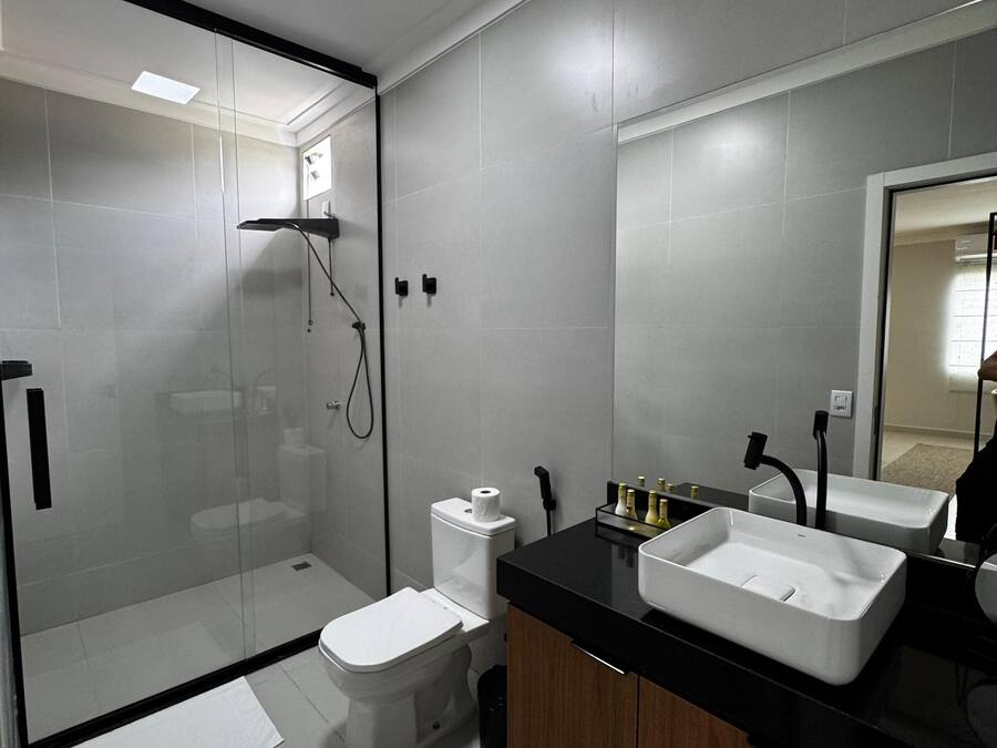 Un baño moderno con ducha envuelta en vidrio, inodoro y dos lavamanos sobre encimera negra.